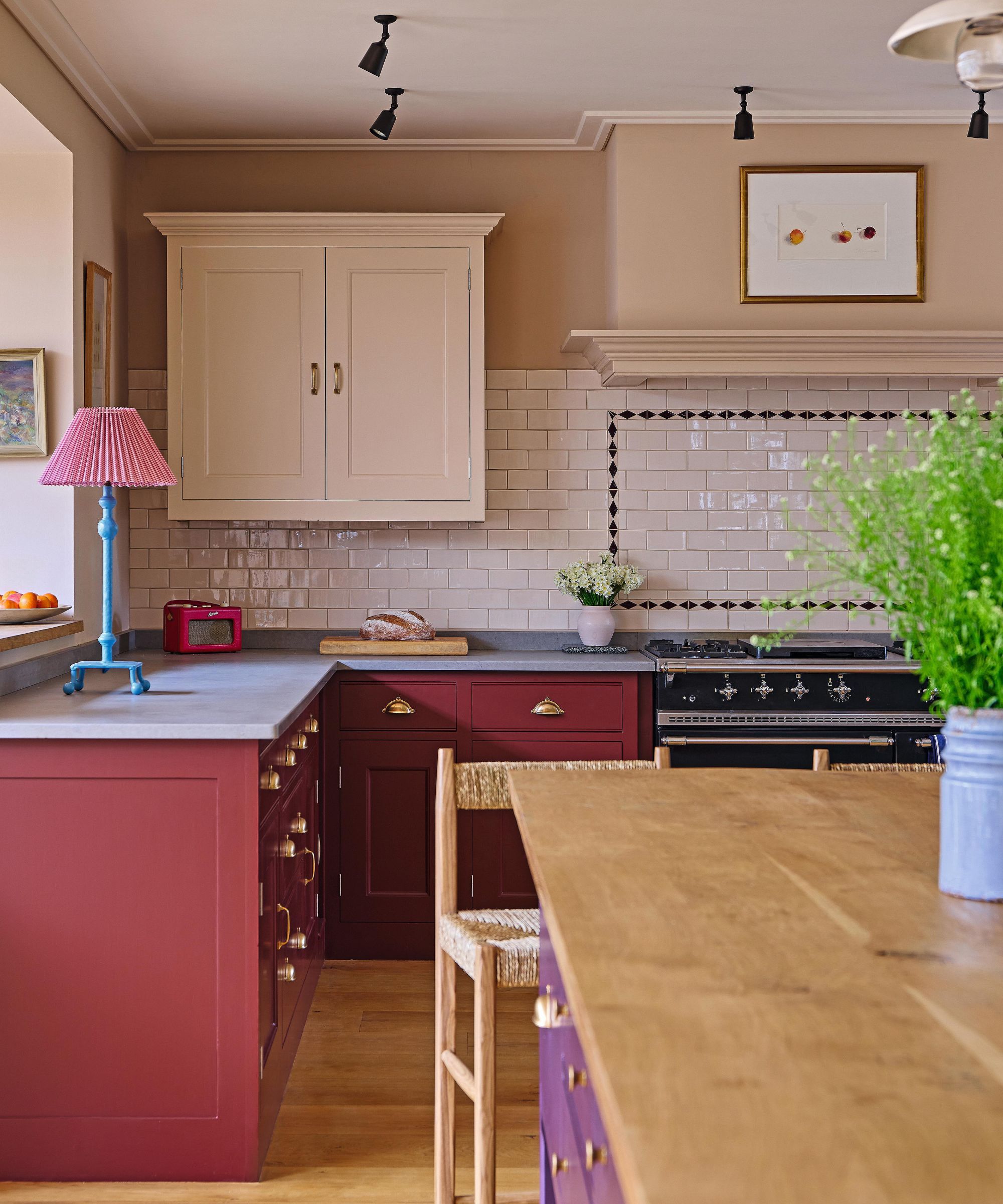 a pale pink english kitchen with tiled backsplash, black cooker, burgundy lower cabinets and a large island