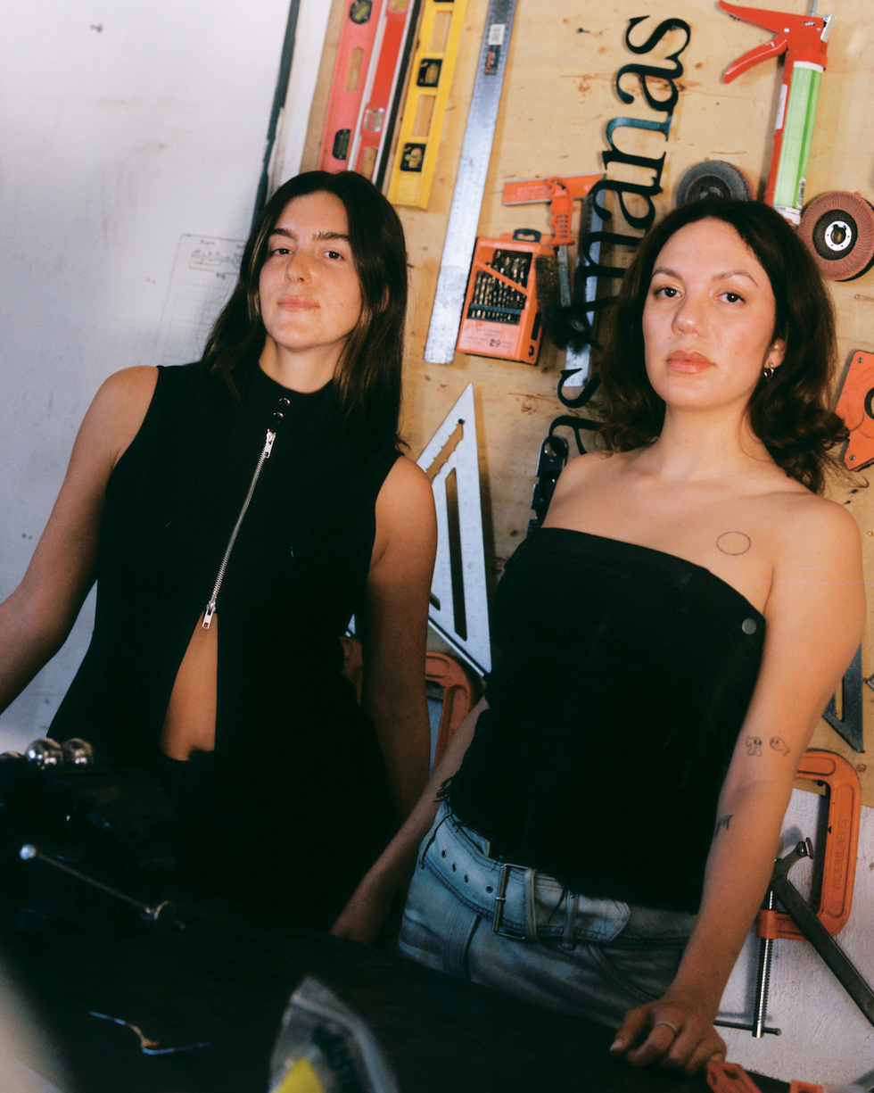 Two individuals posing in a workshop setting with various tools in the background.