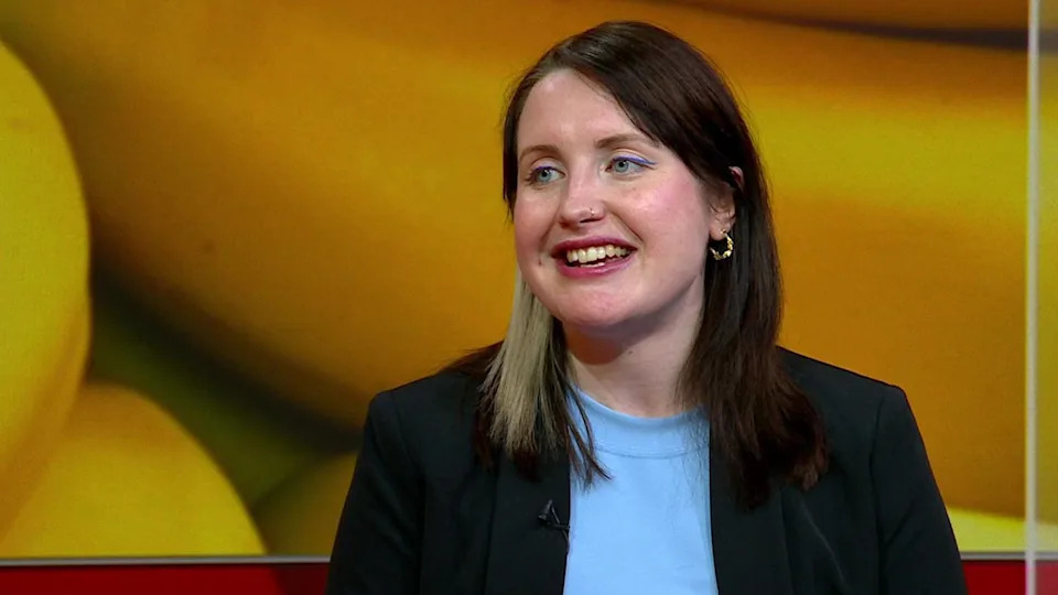 Georgie Oatley sitting on the Look East sofa in the studio. She is looking towards the left at the presenter and has a digital screen of bananas behind her. She is wearing a pale blue T-shirt and a black blazer.