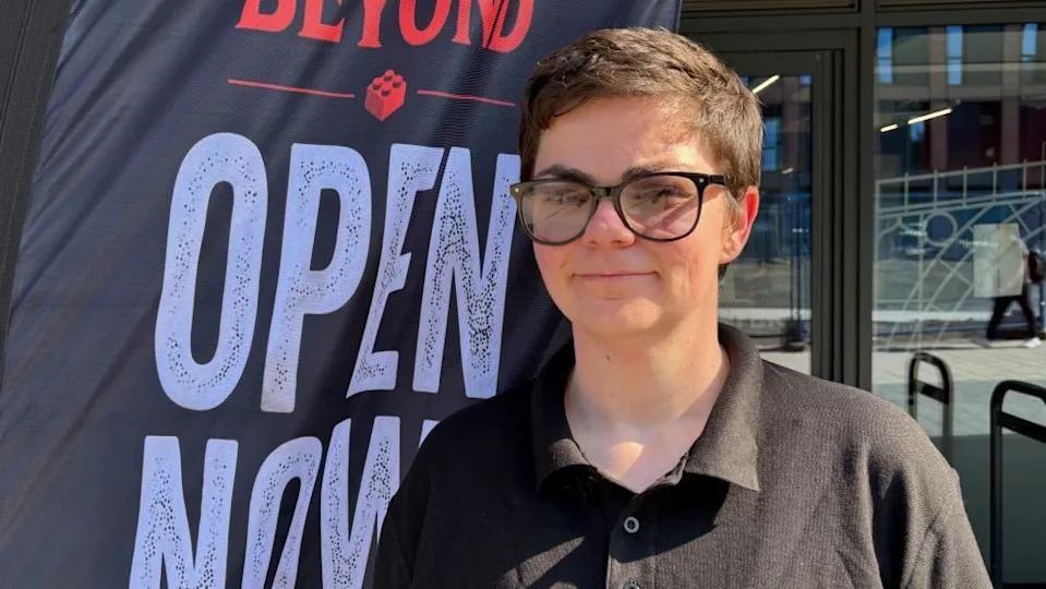 There is a woman with short brown hair and black framed glasses. She is stood next to a banner which says 'Open now' in big white block letters. She is wearing a black polo shirt.