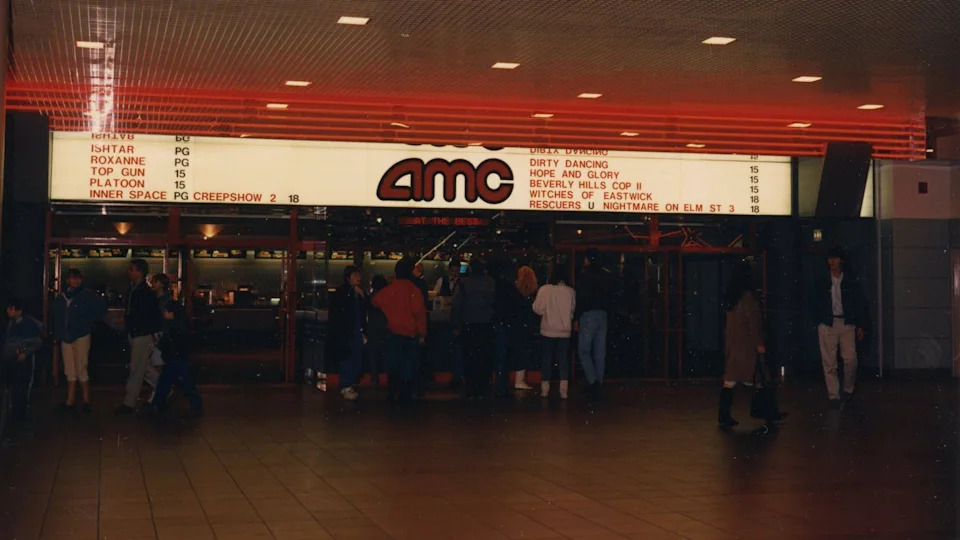 The old cinema which has a lit up board with AMC written in red letters. Some of the films showing are also listed, including Top Gun, Dirty Dancing and Witches of Eastwick. People are gathered below the sign.