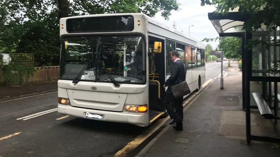 A man in a suit gets on a white single-deck bus at a bus stop in Thames Ditton.