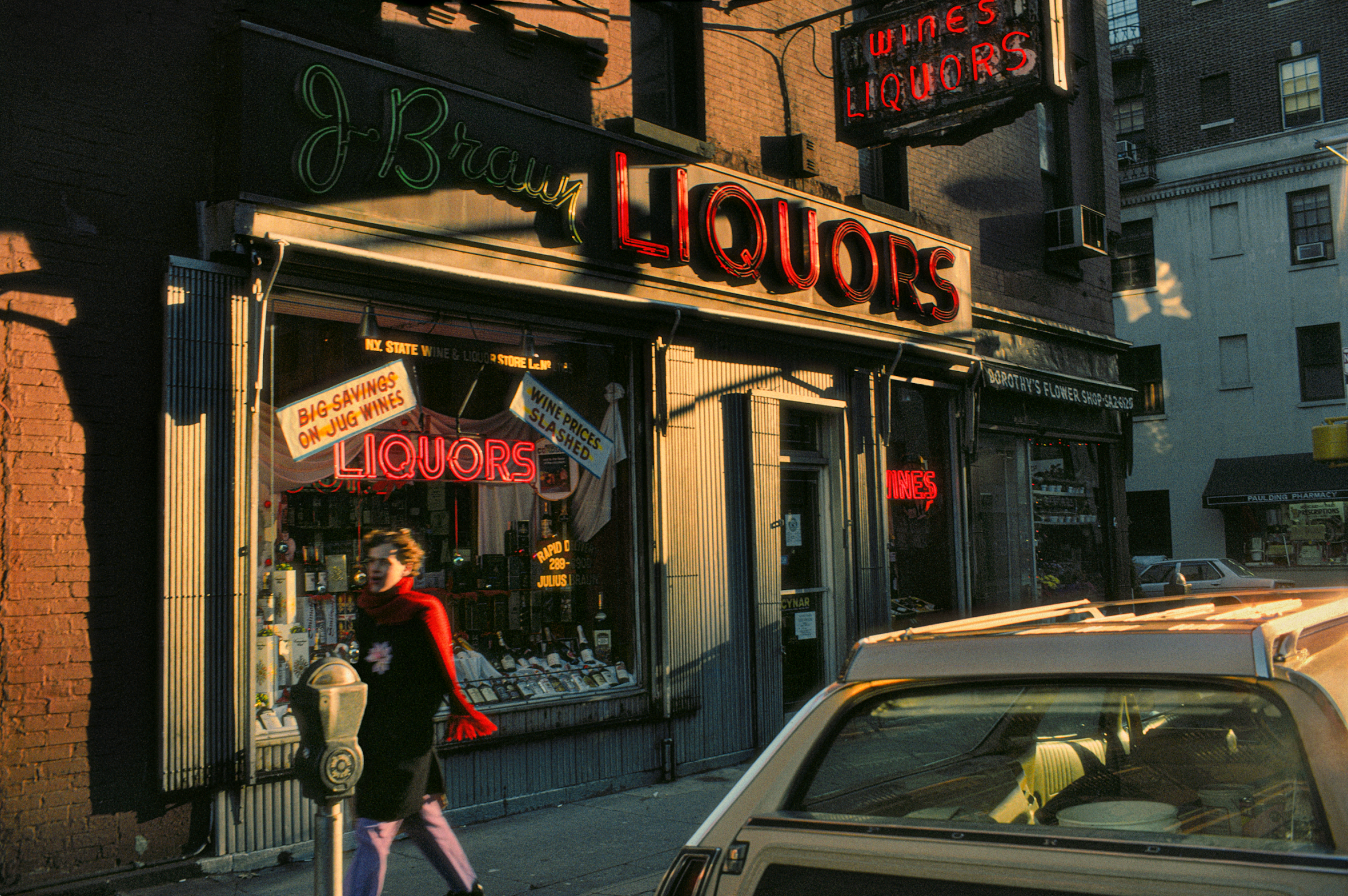 A woman in a red scarf walks past J. Brown Liquors, with neon signs glowing on the store and a parked car in the foreground.