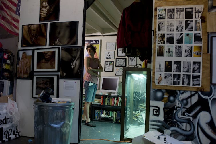 A young man folding his arms is visible through a mirror, which shows a room with walls filled with photographs and other imagery.