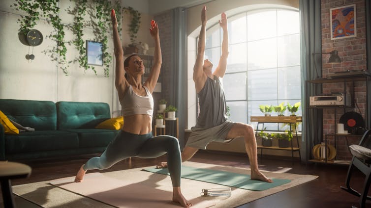 A fit man and woman perform a yoga move in an apartment while the morning sun shines through a window.