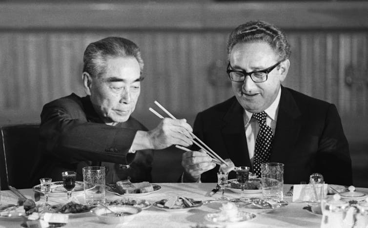 A man uses chopsticks to transfer food to another man's dish