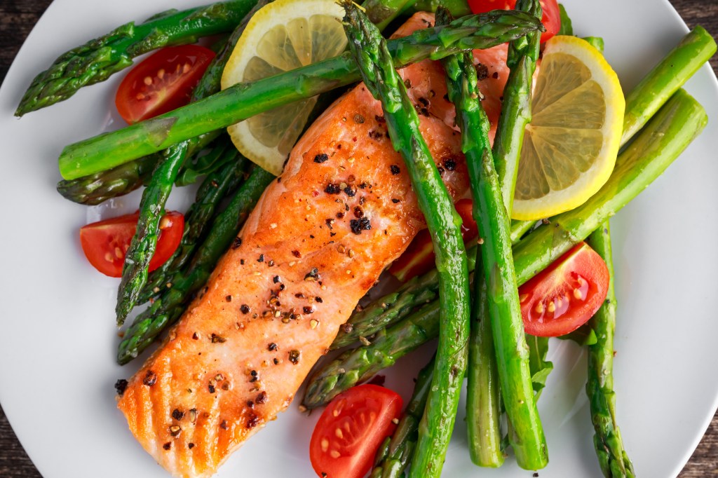 Fried salmon fillet with asparagus, tomatoes, and lemon slices on a white plate.