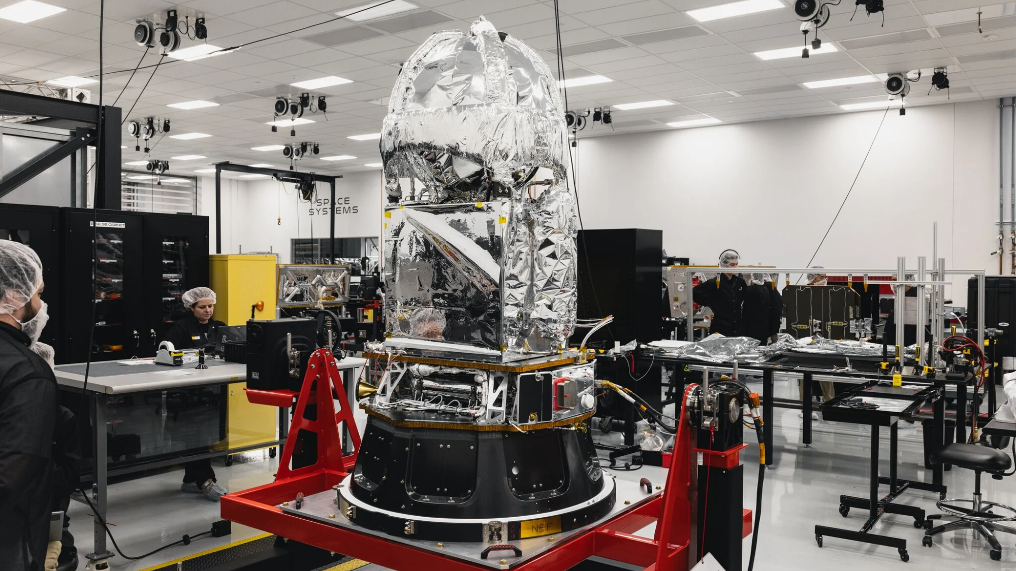 a large tapered cylindrical space probe sits, wide side down, on a red frame. It is black on the bottom and covered in silver foil on the top. In a white room with technicians in hair nets.
