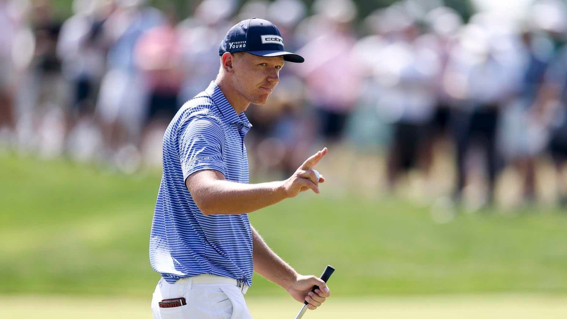 A golfer in a blue striped shirt and cap holds a putter and gestures with two fingers raised, while standing on a golf course with a blurred crowd in the background.