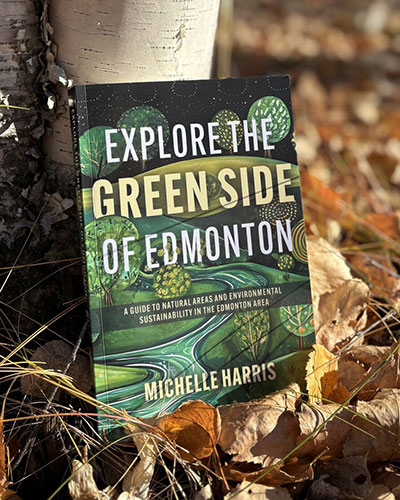 Book titled “Explore the Green Side of Edmonton” by Michelle Harris placed against a tree among fallen autumn leaves.