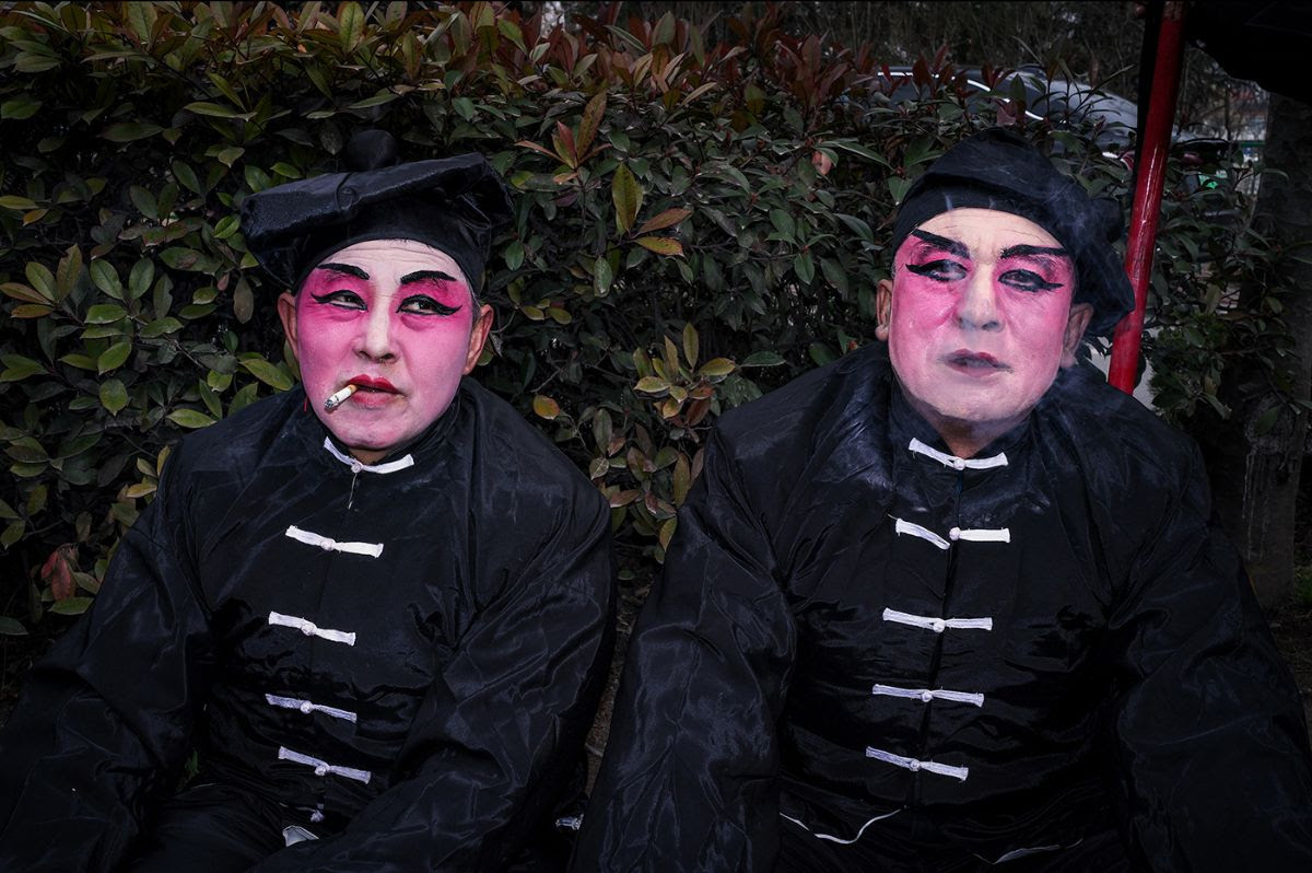 Two performers with vivid pink face makeup and dramatic black eye markings wear matching black traditional costumes with white toggle closures against a backdrop of dark foliage, one smoking a cigarette.