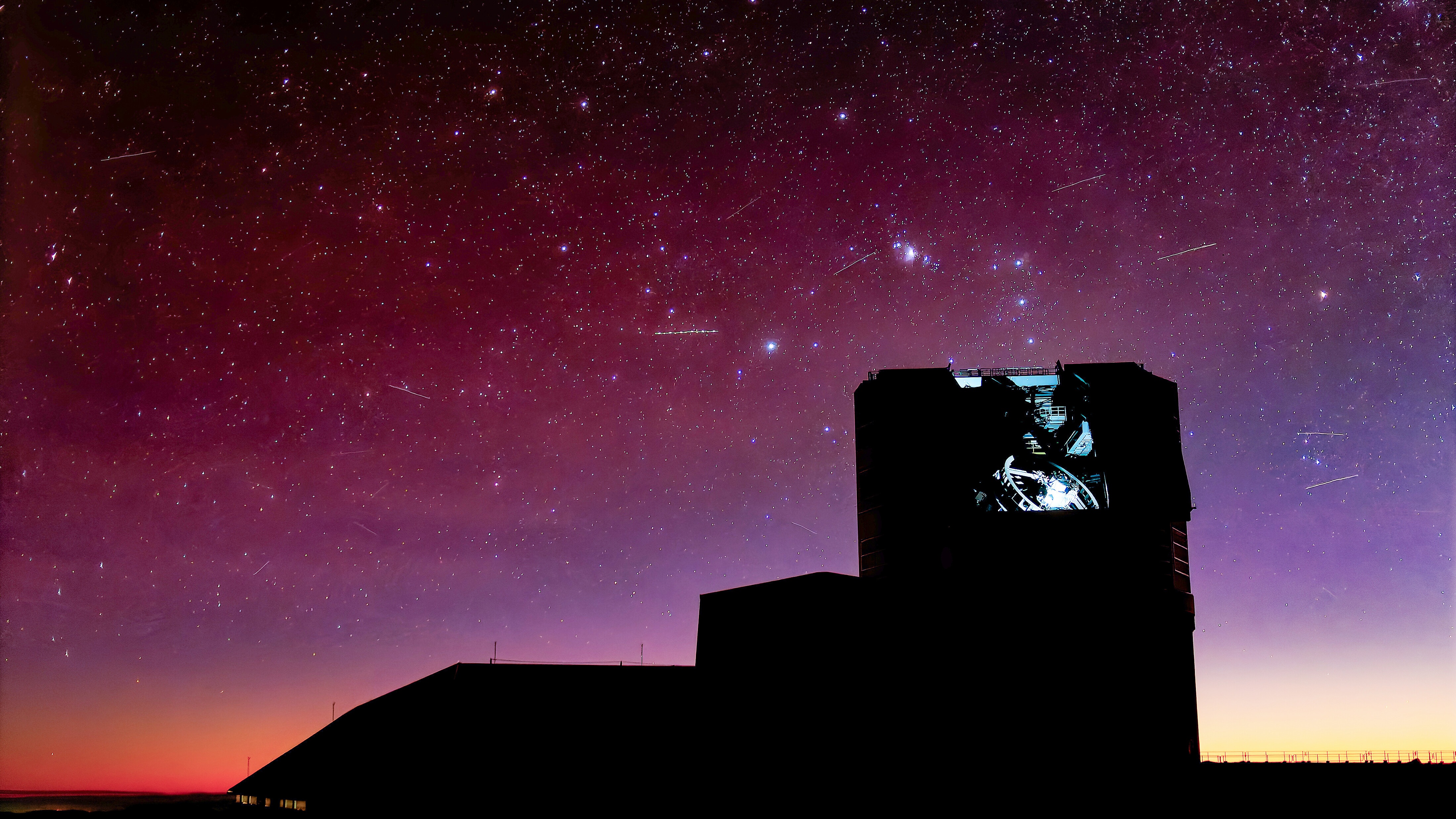 The silhouette of an observatory under a pinkish sky with lots of stars and a few streaks.