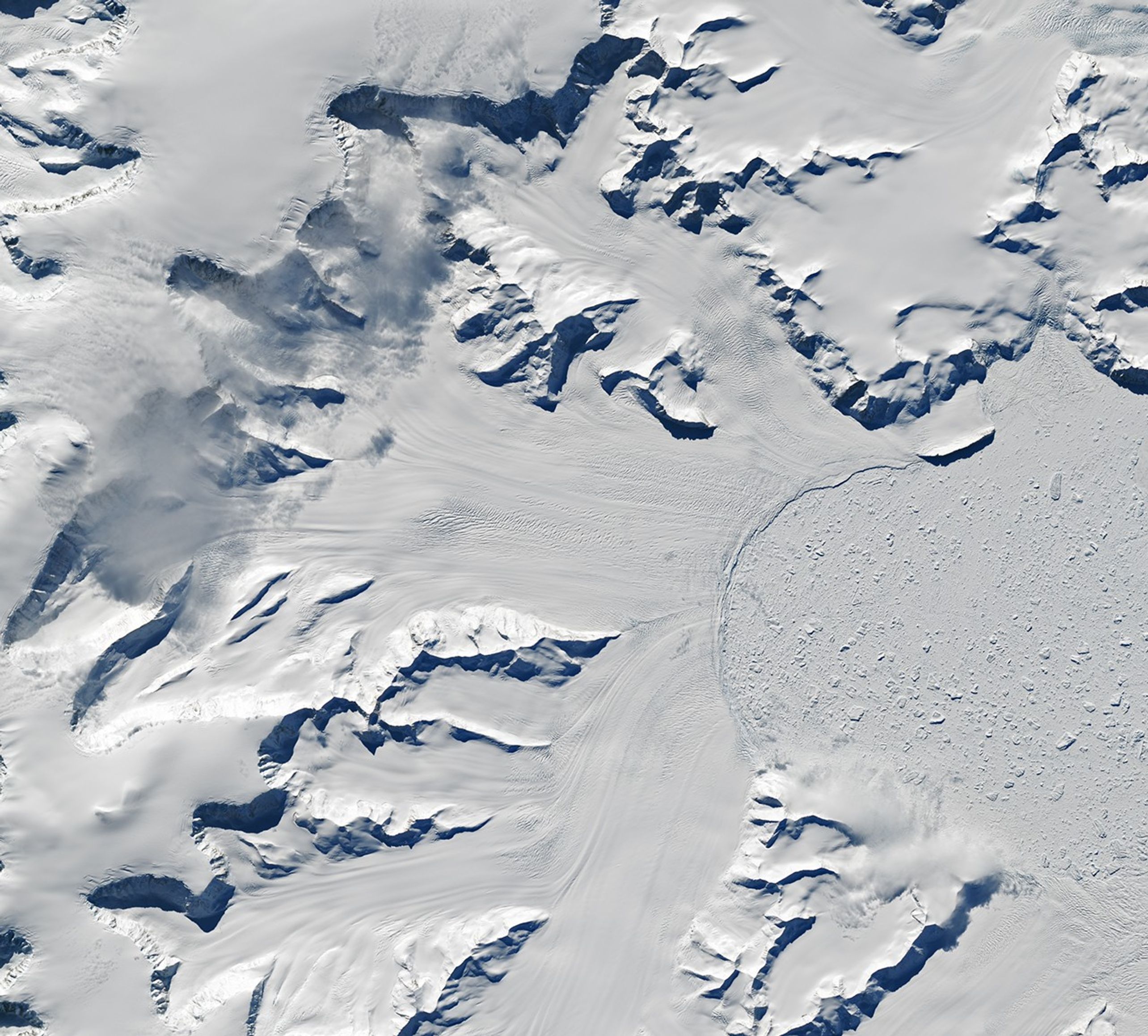 Glaciers flow from cliffs on the left and bottom sides of the image to a frozen bay on the right where chunks of sea ice are visible.