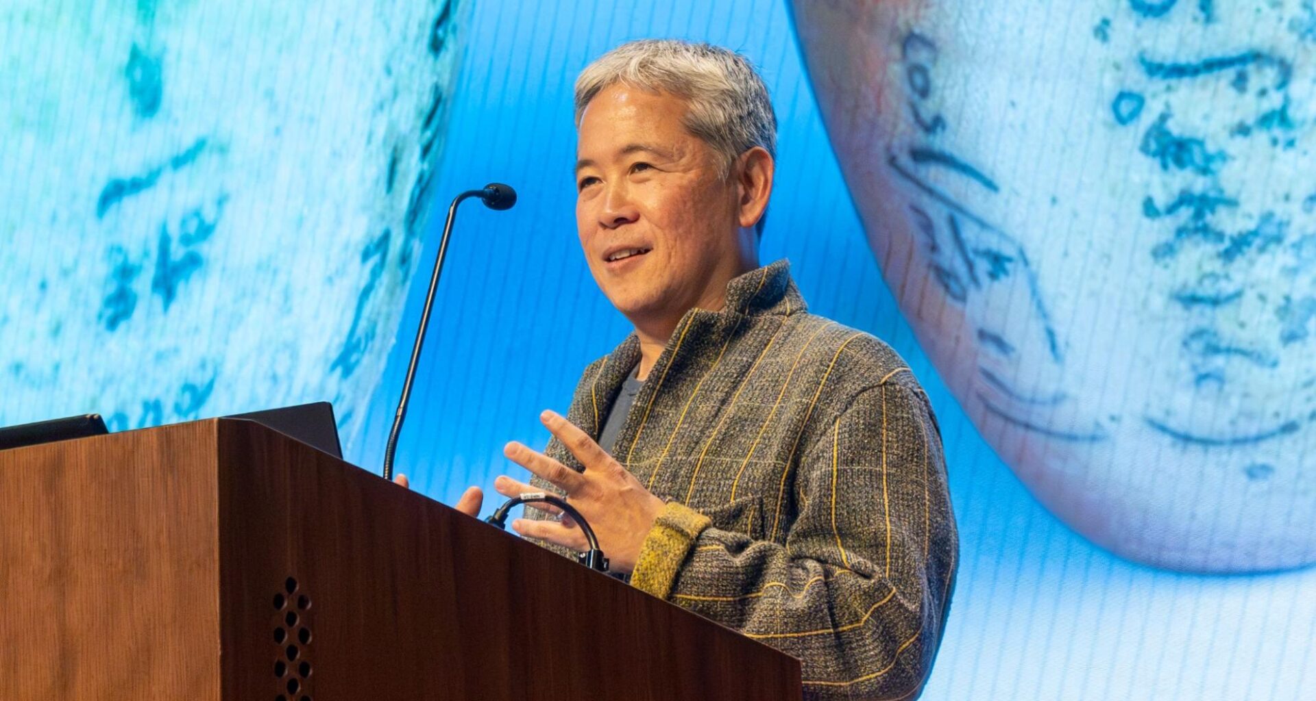 Hideo Mabuchi speaking on a lectern, with a photo of ceramics projected in the background