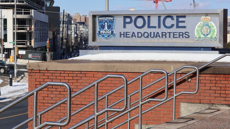 A steel sign that says Police Headquarters with a blue and white logo with a lighthouse and Halifax Regional Police to the eft and another logo with a crown to the right stands atop a red brick wall covered in snow. Below steel railings descend down a concrete staircase on the front of a building.