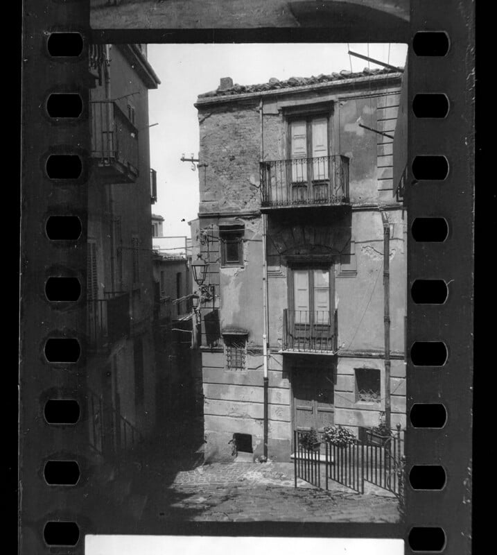 Black and white photo framed by film strip edges, showing an old European street with a weathered building, balconies, shuttered windows, cobblestones, and iron railings.