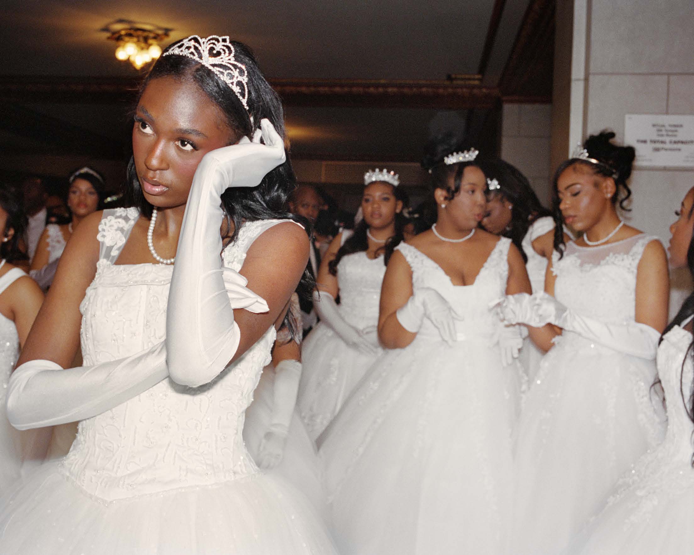 A young Black woman in a white ballgown, tiara and long white gloves looks directly at the camera with a composed, watchful expression, surrounded by other similarly dressed women in a formal interior.