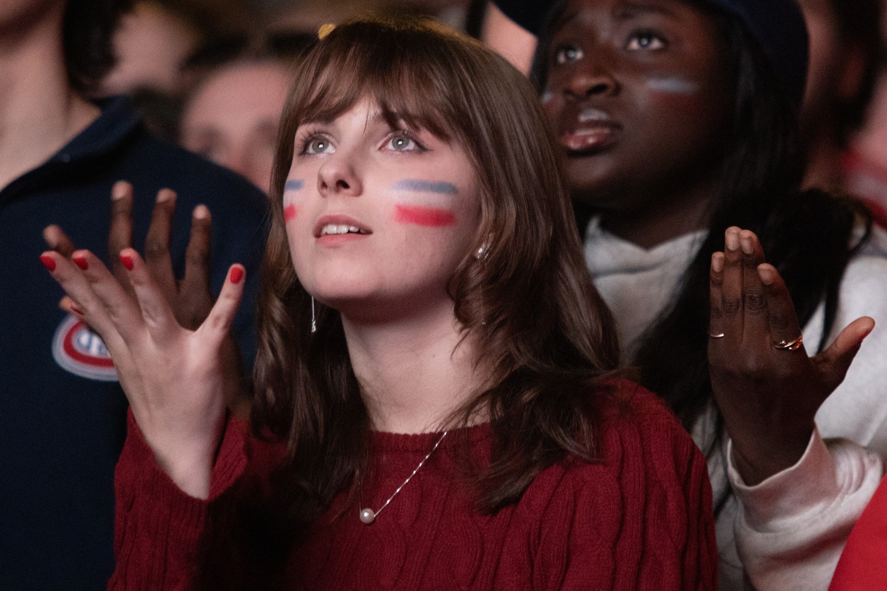 A young woman with a disbelieving stare holds out her hand in a crowd as she looks up. Her cheeks are painted with the colours of the Montreal Canadiens.