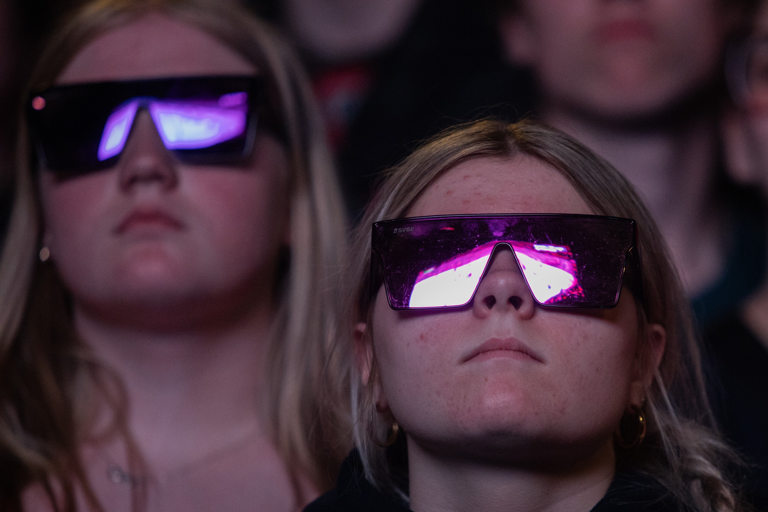 Two sombre women with sunglasses on, with a screen reflected in their glasses. The screen is showing the rink at a Canadiens game.