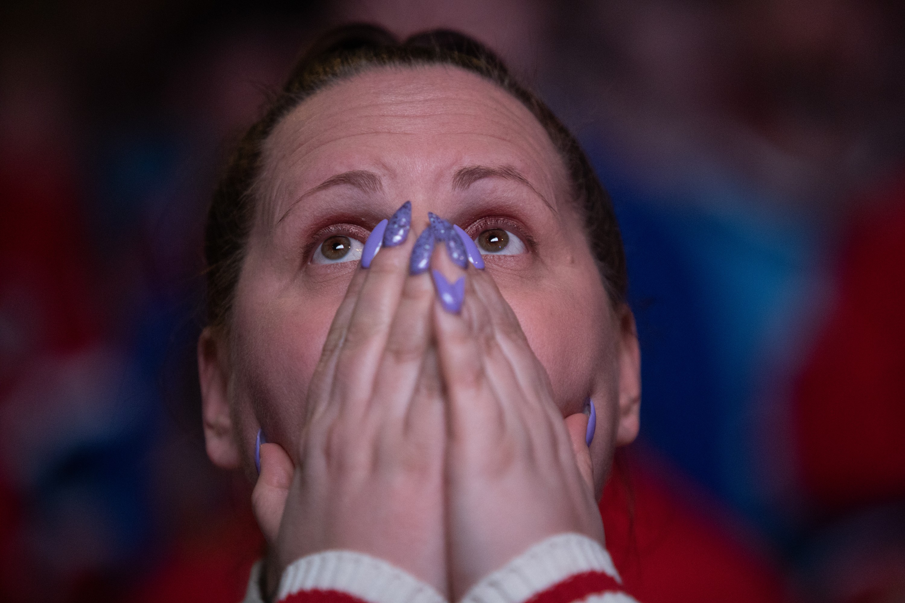A woman holds her hands up to her face, covering her mouth, as her eyes are wide while watching an unseen screen.