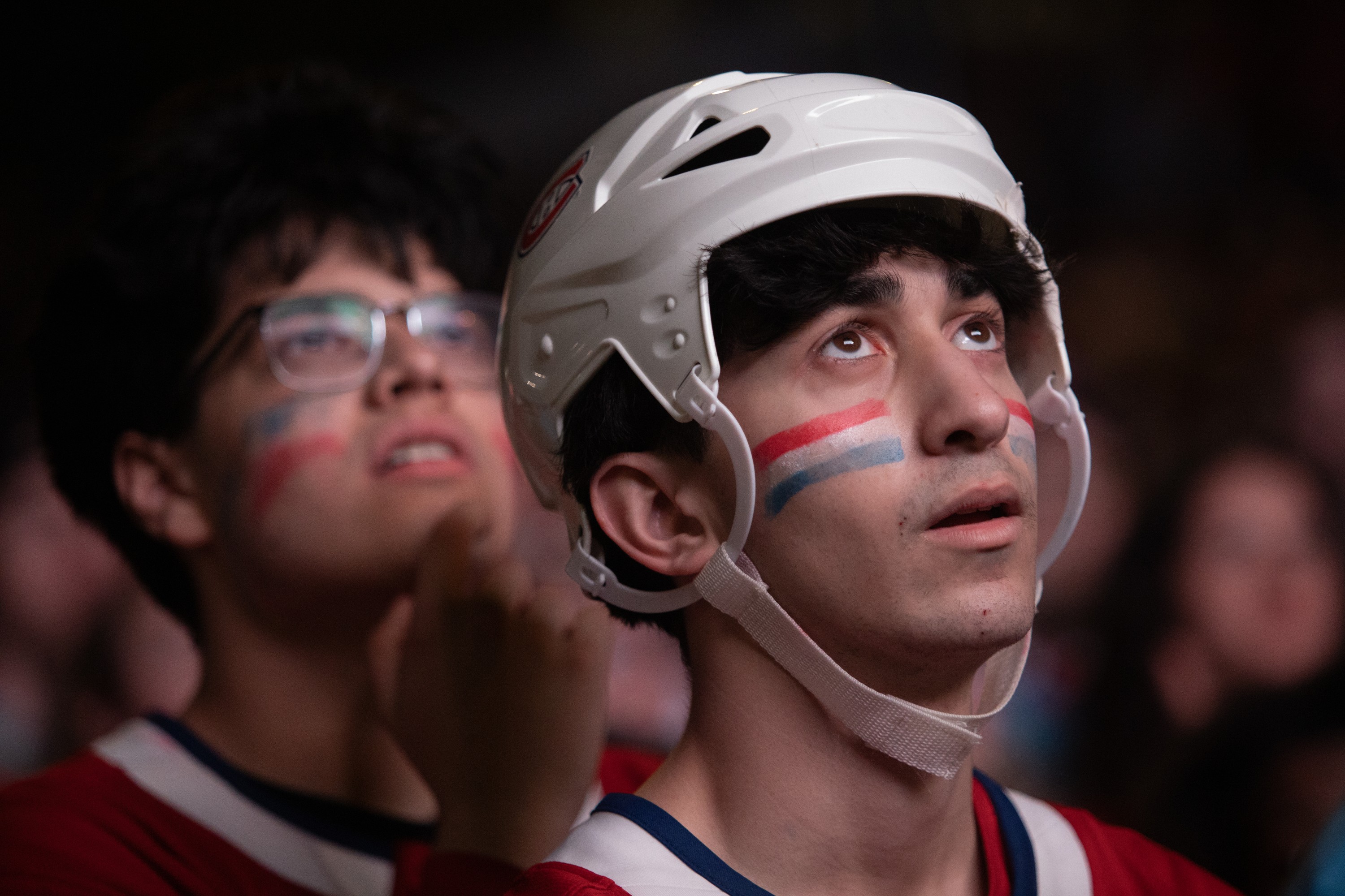 Two Montreal Canadiens fans don't look happy as they watch an unseen screen. Both have the team colours painted on their cheeks, and the one in the foreground is wearing a hockey helmet.