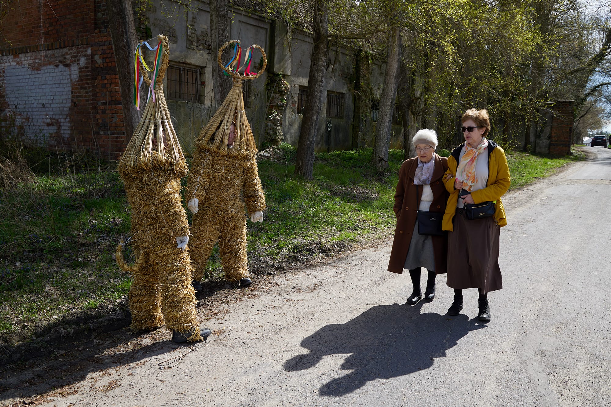 A candid street photograph by Janusz Jurek of two elderly women walking by costumed and masked figures during a folk festival