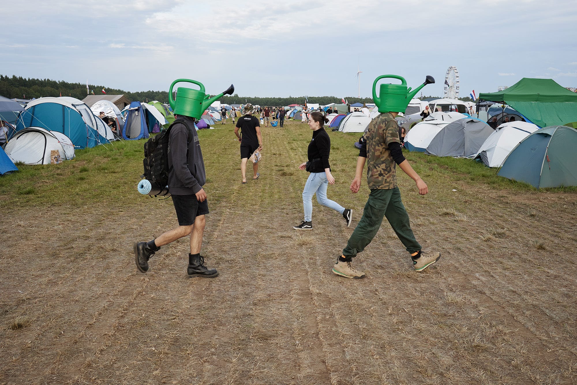 A candid street photograph by Janusz Jurek of two young men walking across a festival grounds with green watering cans on their heads