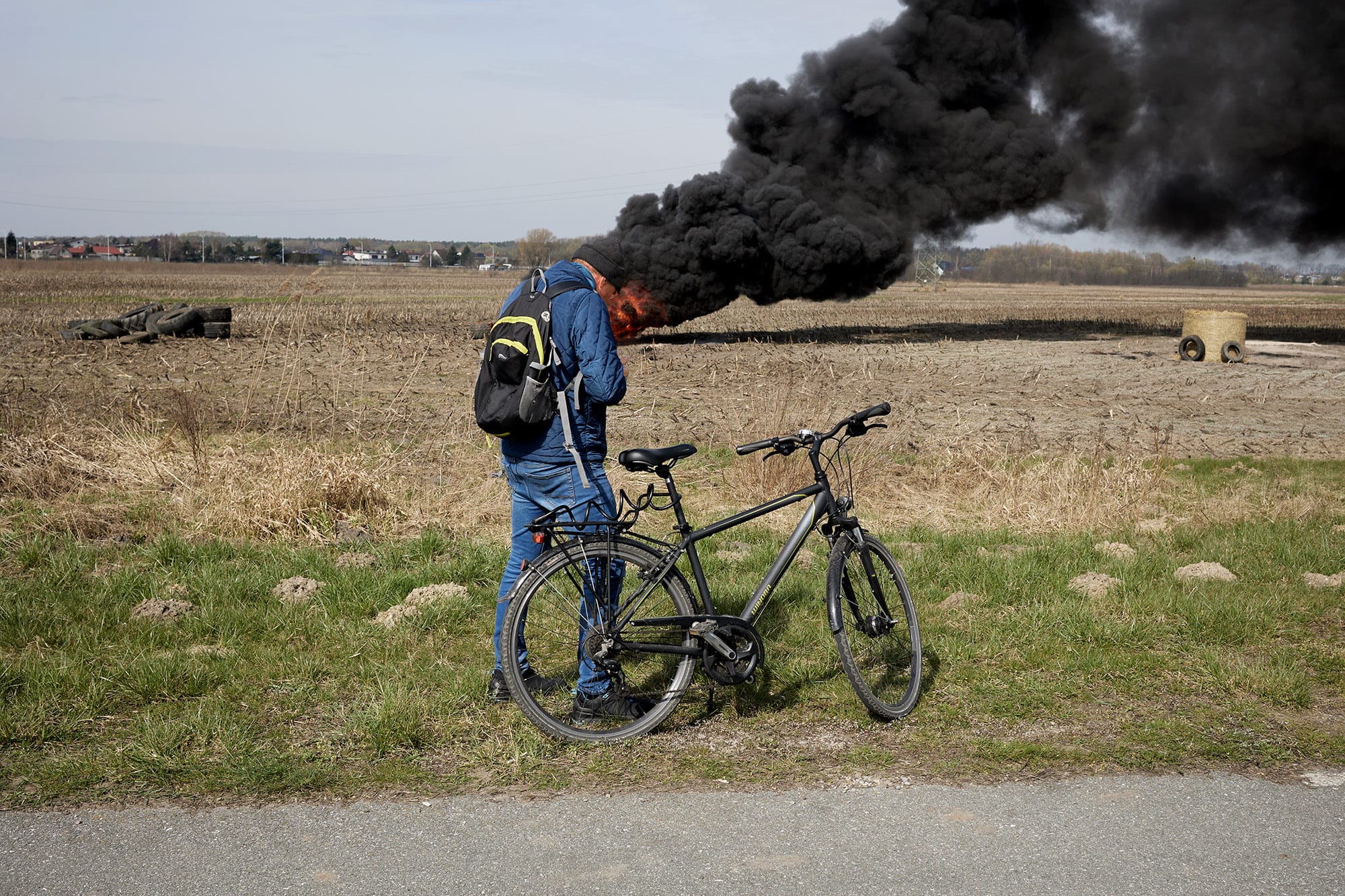 A candid street photograph by Janusz Jurek of a man stopped on a bicycle near a large fire in a field, and the smoke looks as though it's coming out of his head