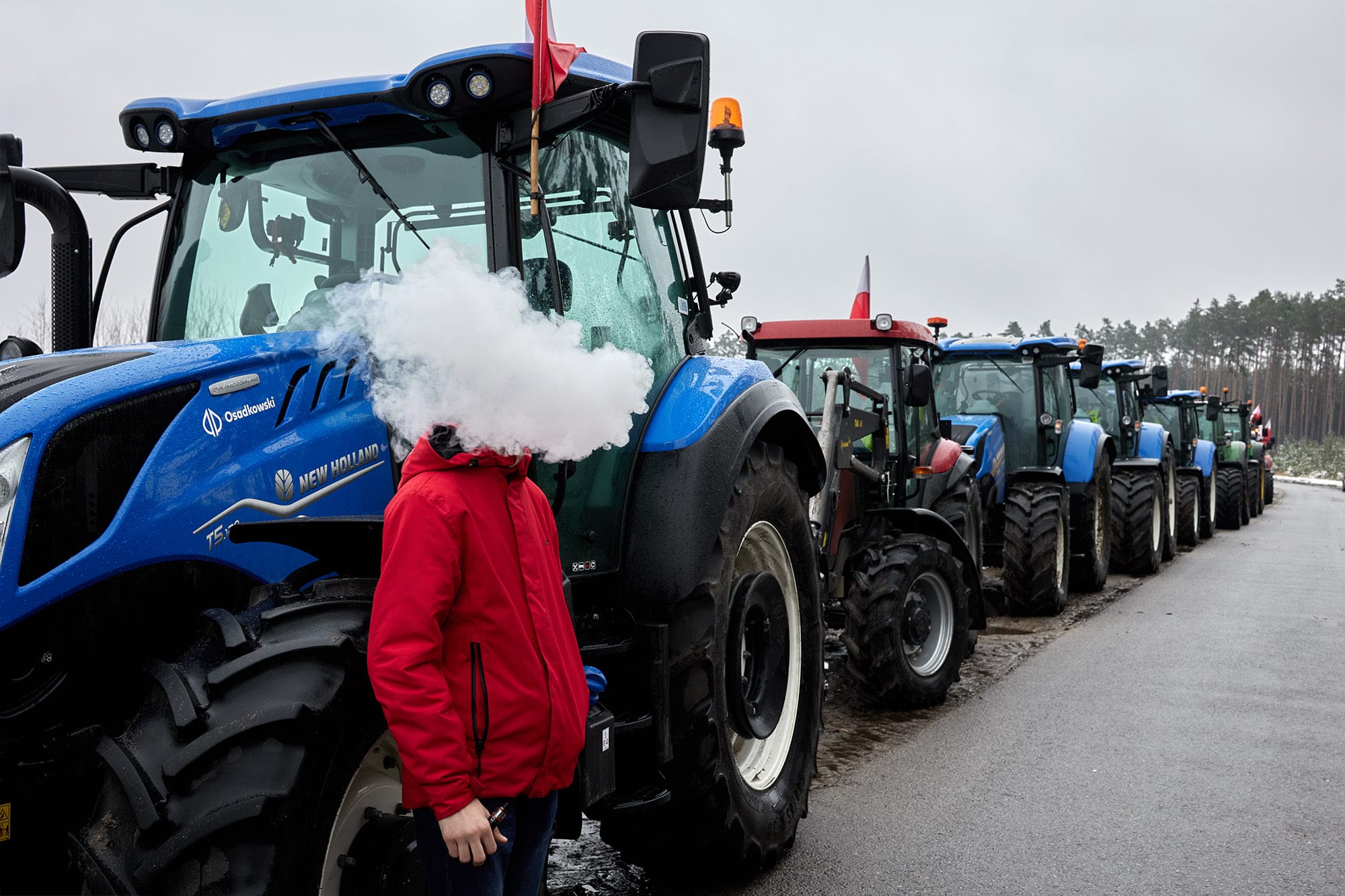 A candid street photograph by Janusz Jurek of a man standing in front of a blue tractor with smoke covering his face