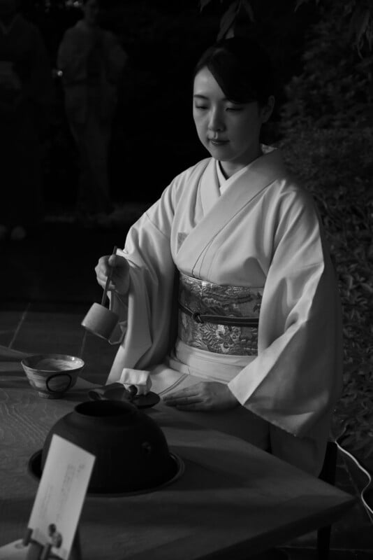 A woman in traditional Japanese attire performs a tea ceremony, gracefully holding a ladle over a tea set on a wooden table, with foliage and another person blurred in the background.
