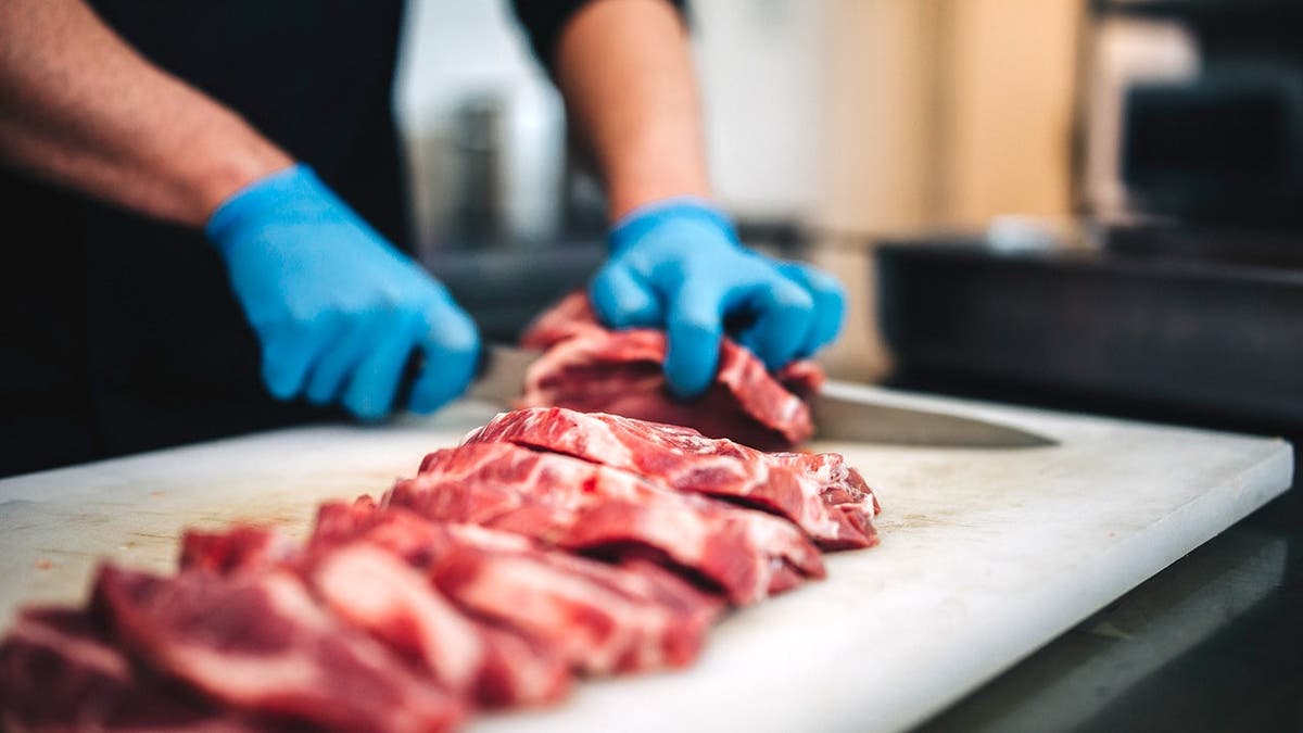 A man wearing disposable gloves cuts a piece of beef with a knive.