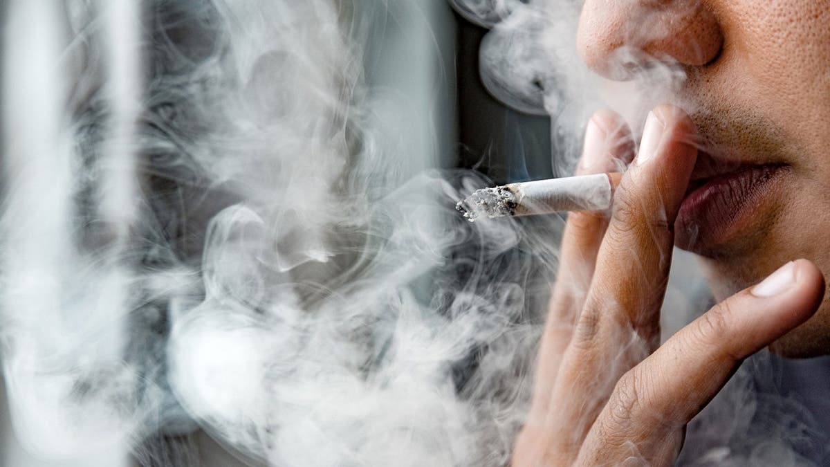 A man takes a drag from a cigarette while smoke surrounds him.