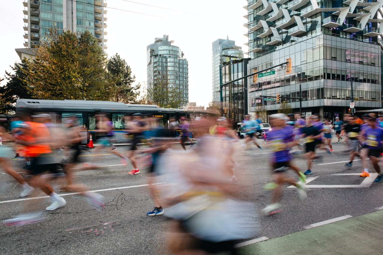 Motion-blurred runners race through a downtown Vancouver intersection during a marathon, passing in front of a TransLink bus as glass high-rise towers and city streets frame the background