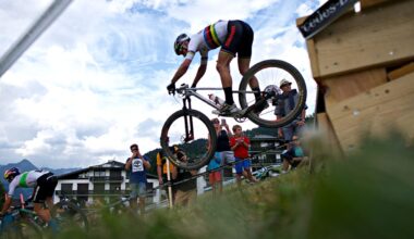A mountain biker performing a jump over a wooden ramp during a race, with spectators gathered on the sidelines watching and taking photos. The scene is set against a backdrop of cloudy skies and mountains, capturing the excitement of the event.