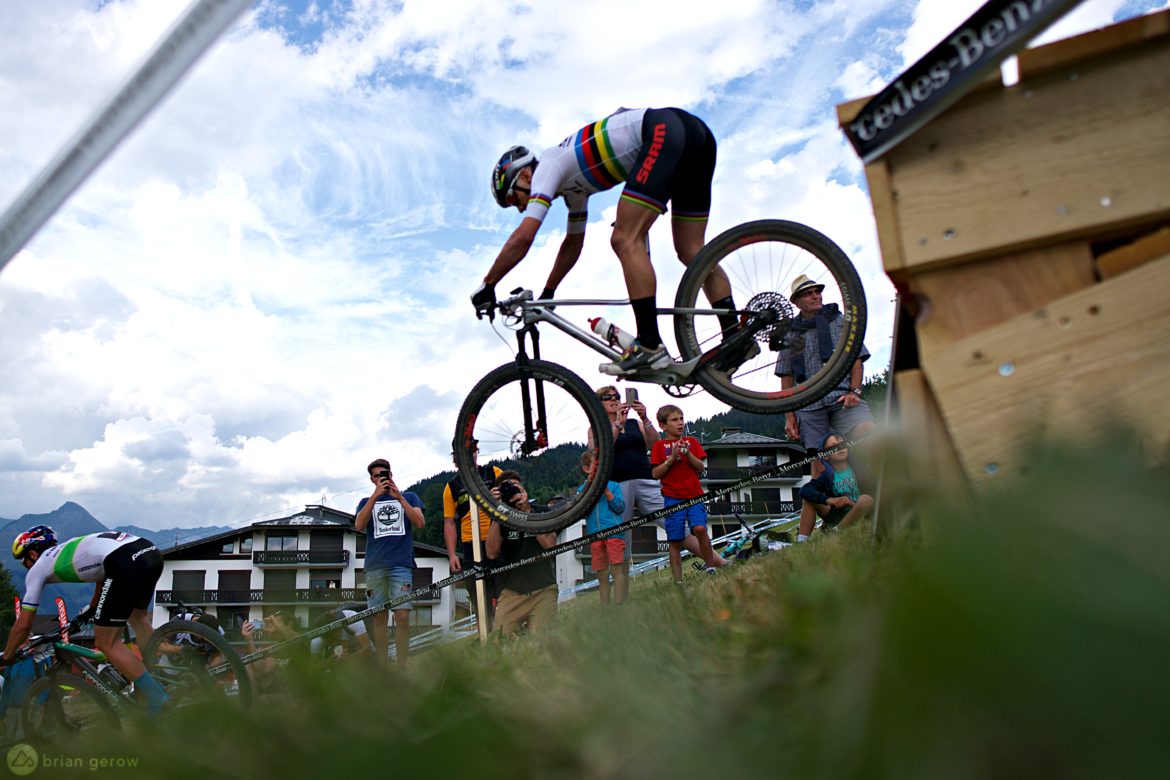 A mountain biker performing a jump over a wooden ramp during a race, with spectators gathered on the sidelines watching and taking photos. The scene is set against a backdrop of cloudy skies and mountains, capturing the excitement of the event.