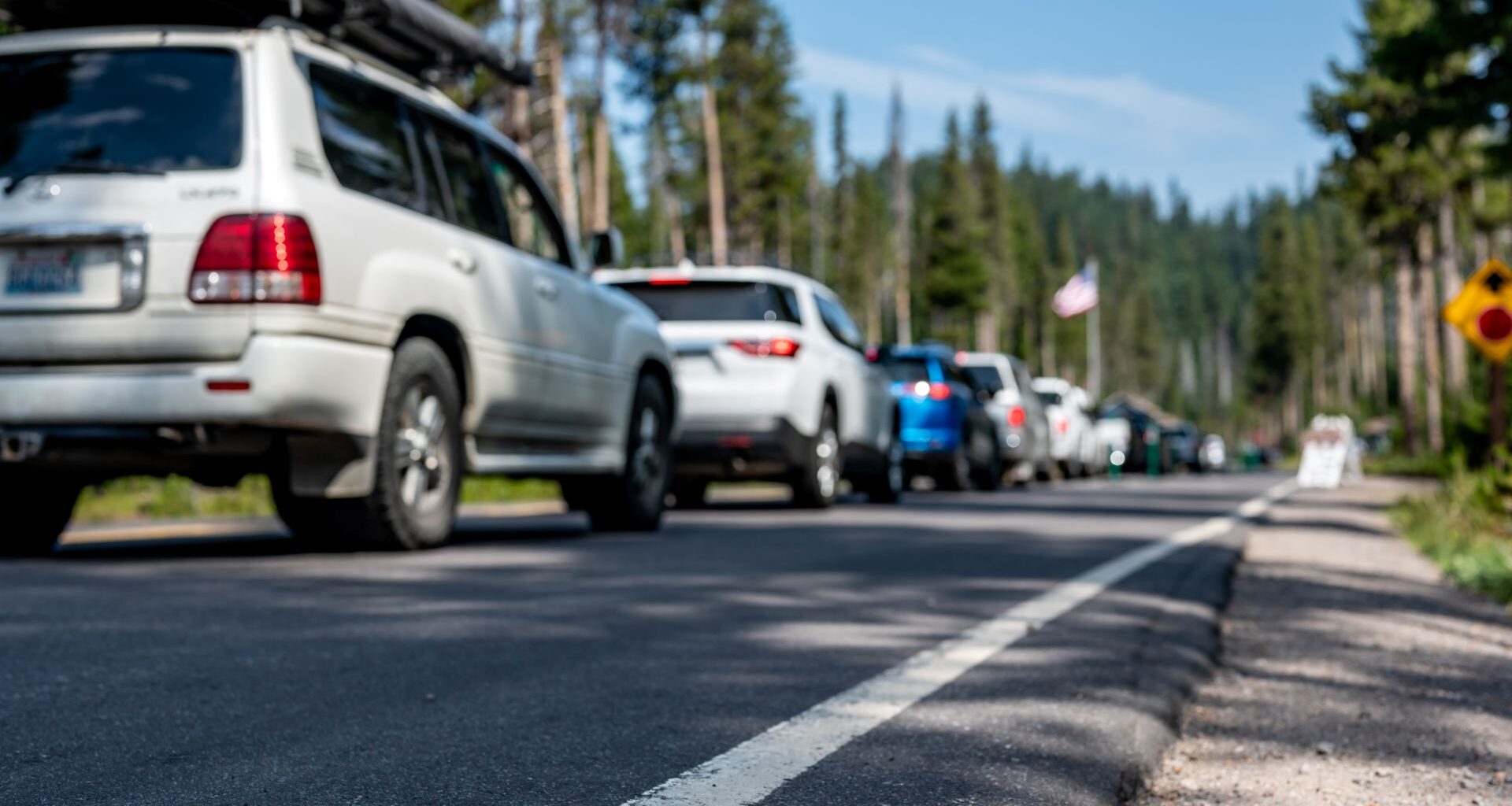 A line of cars waiting on a road with trees and a clear blue sky in the background.