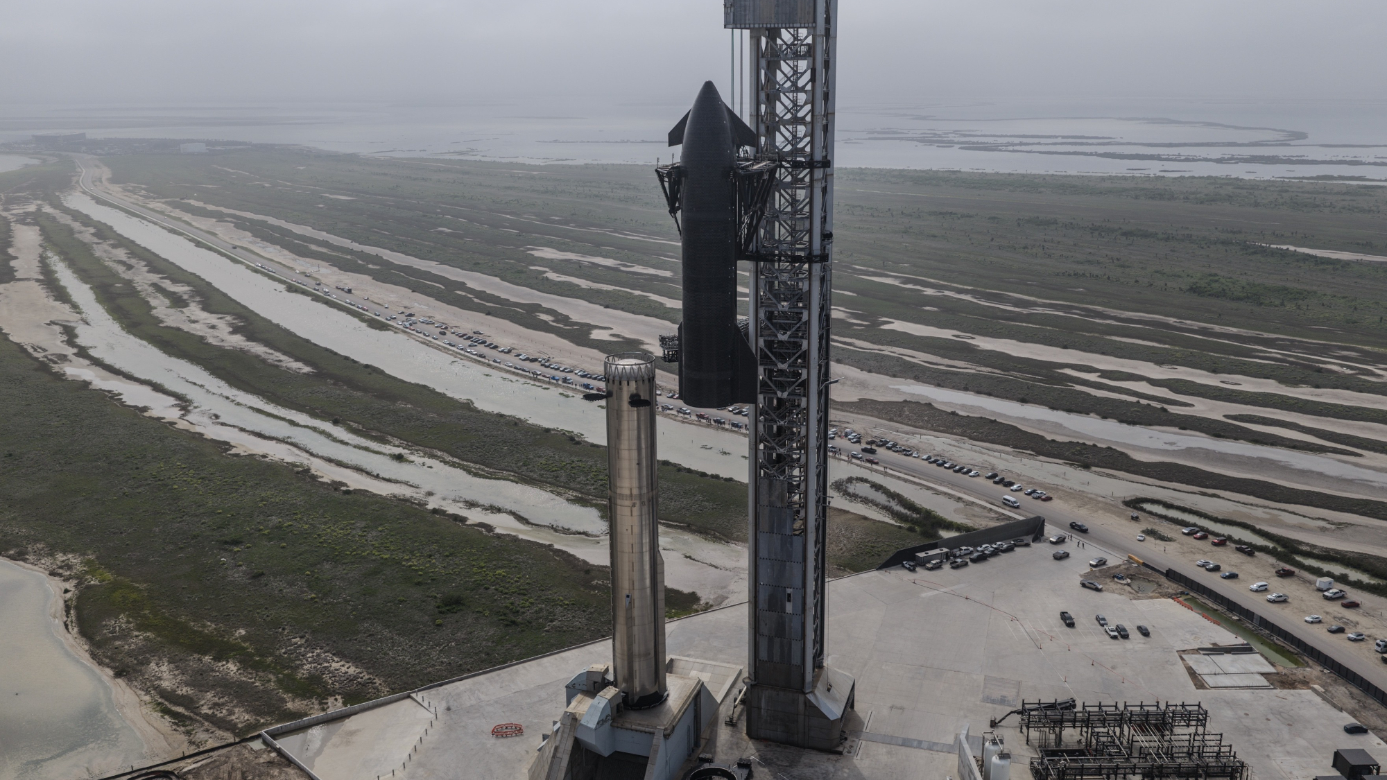 a giant grey tower lifts a black spaceship to stack on a chrome rocket booster amid flat coastal water lands near the shore.
