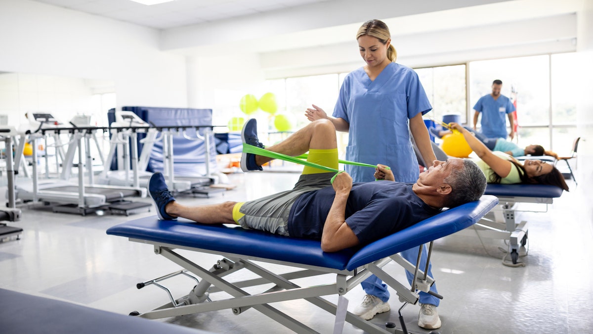 Mature Latin American man doing physical therapy exercises with a stretch band assisted by therapist