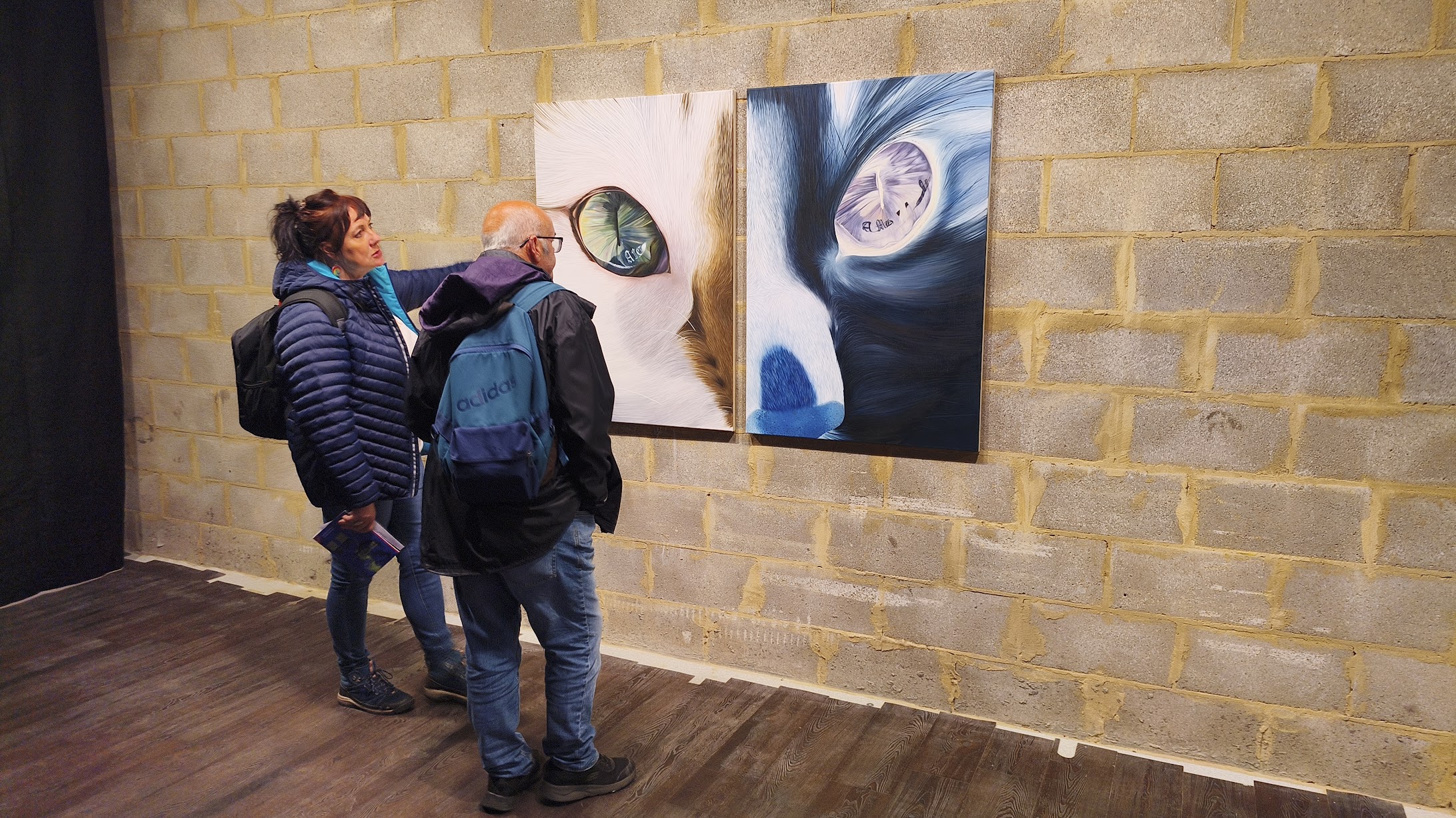 wo people stand in an industrial-style gallery with cinder block walls, viewing a large diptych painting of a cat's eyes.