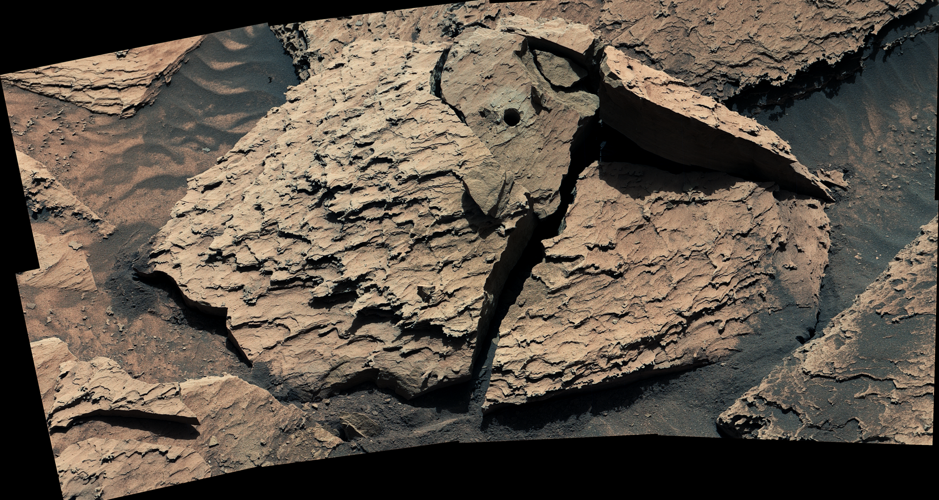 A large, reddish-brown rock as seen by NASA's Curiosity Mars rover. The rock is broken into several pieces. A drill hole is visible in the top middle piece.