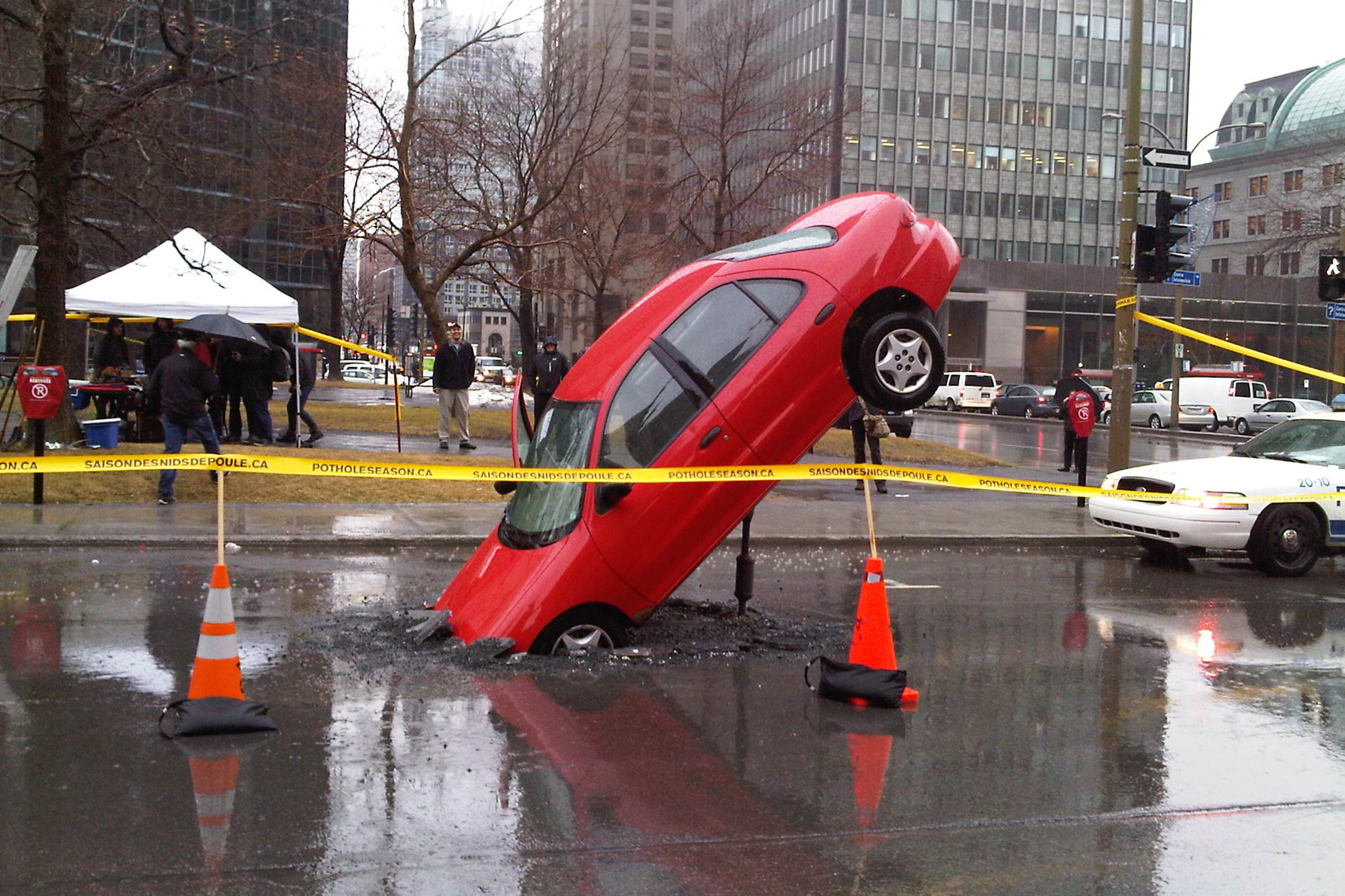A car is pitched over nearly vertical with its front end in a pothole in a downtown street.