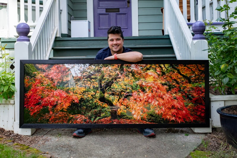 A person sits on porch steps, smiling and holding a large framed photograph of a vibrant autumn tree with red and orange leaves. Behind them is a purple door and white railing with purple accents.