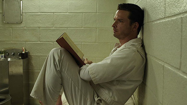 Daniel Holden in white prison jumpsuit sitting on bed with book