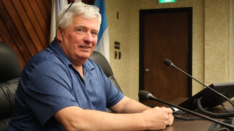 A white man with silver hair, glasses sitting on his head, and wearing a blue shirt-sleeved shirt stands sits in a black office chair behind a long table. There are two microphones and a laptop on the table next to him and a door in the background.