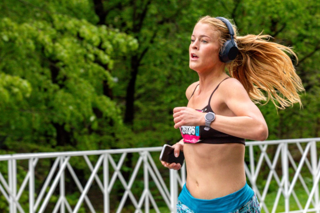 A female runner in black sports bra and blue shorts, wearing headphones and a smartwatch, runs with her hair flying, carrying a phone.