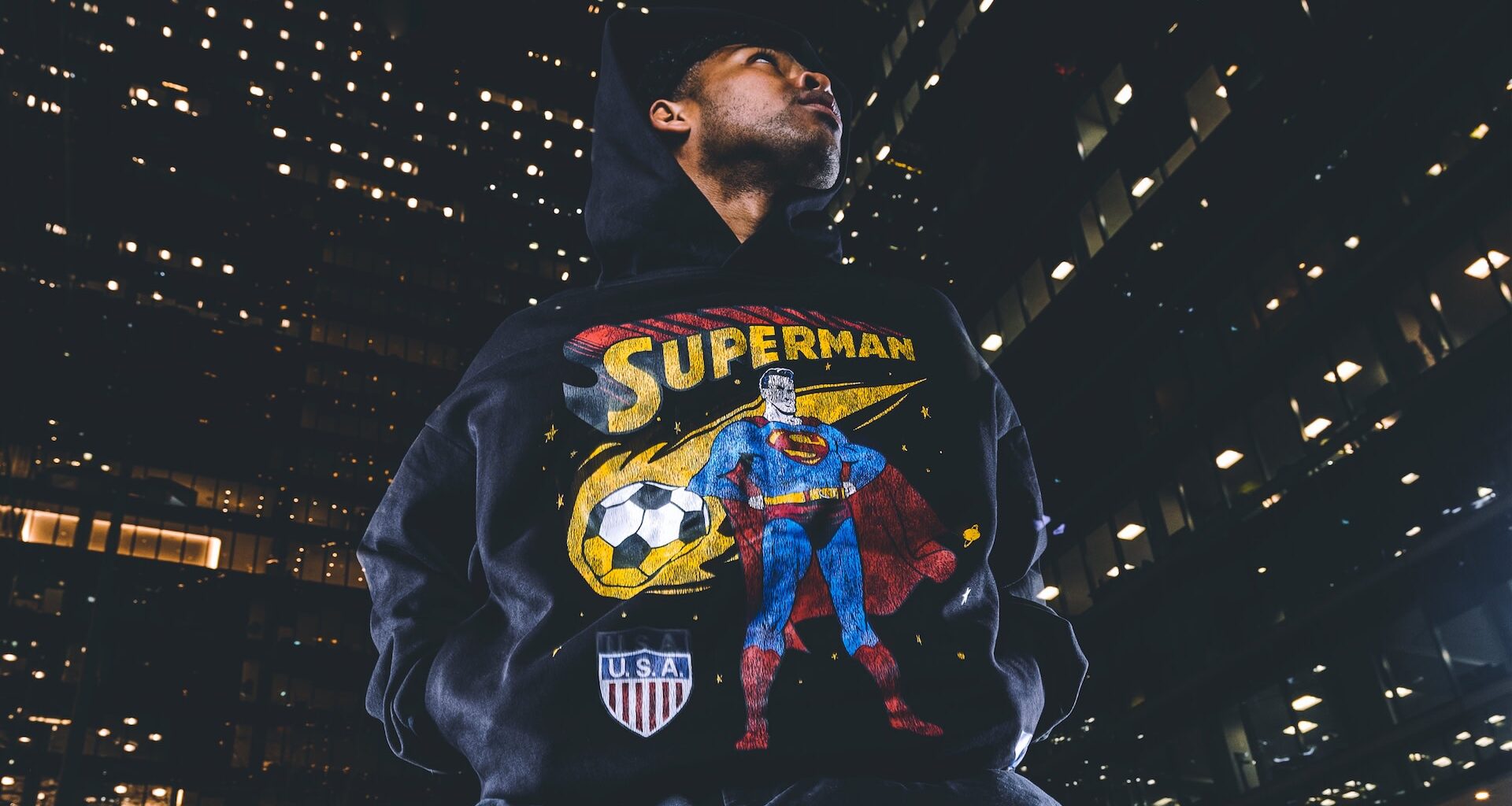 A person wearing a hoodie with a Superman design, featuring a soccer ball and a USA shield, stands against a backdrop of city lights.
