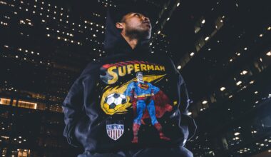 A person wearing a hoodie with a Superman design, featuring a soccer ball and a USA shield, stands against a backdrop of city lights.