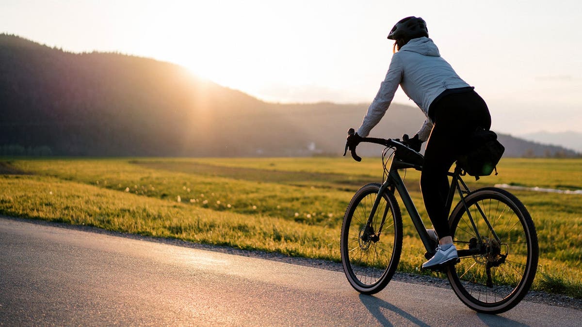 A woman rides a bicycle as the sun rises over a mountain ahead of her.