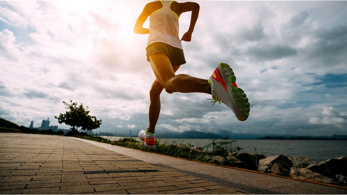 A woman runs on a paved surface toward a bright sunlight and clouds.