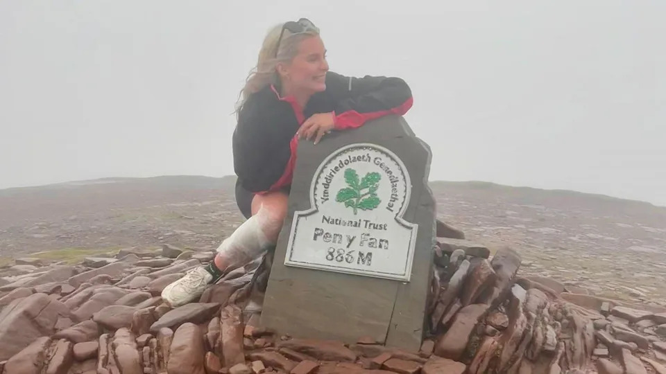 A woman crouched in front of the Pen-y-Fan sign on the top of the summit. She's wearing shorts, a tacked and trainers with a white bandage on her right leg. She's surrounded by brown slab stones.